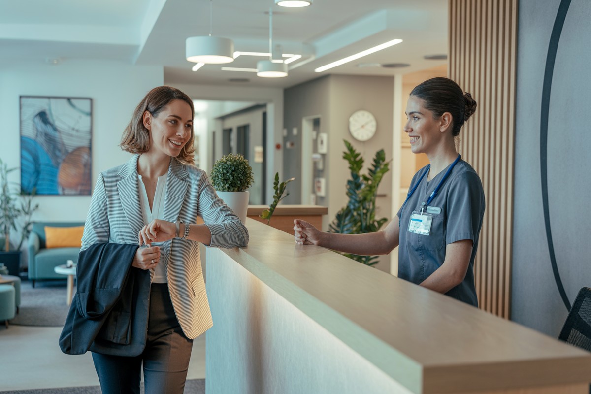 Pracownica w recepcji kliniki medycyny pracy sprawdza zegarek i rozmawia z recepcjonistką