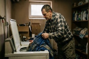 Zmęczony mechanik wkładający brudny, roboczy kombinezon do pralki w domowej piwnicy.